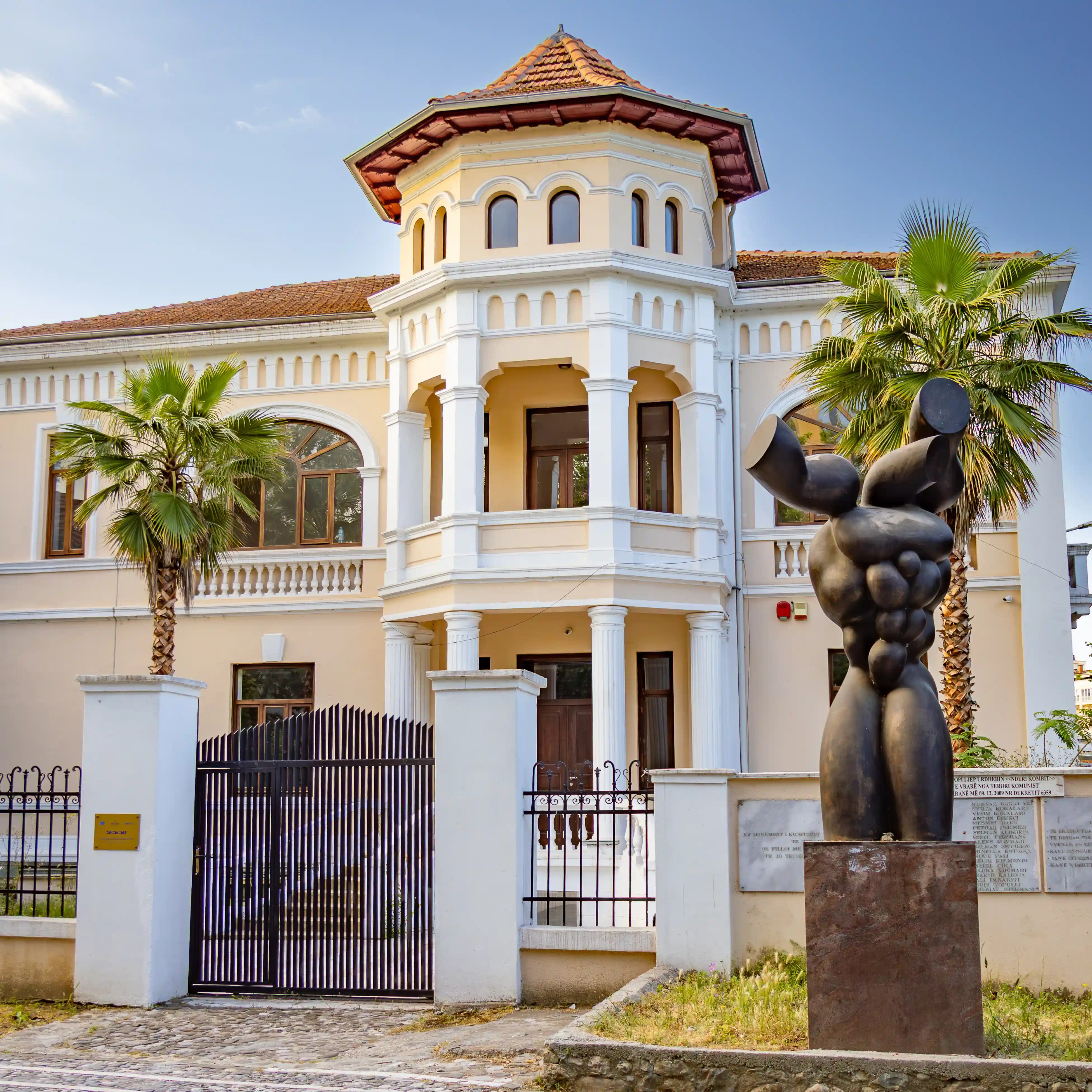 A cream-colored historic villa with a tower-like entrance, palm trees, and a large modern sculpture in front of the building.