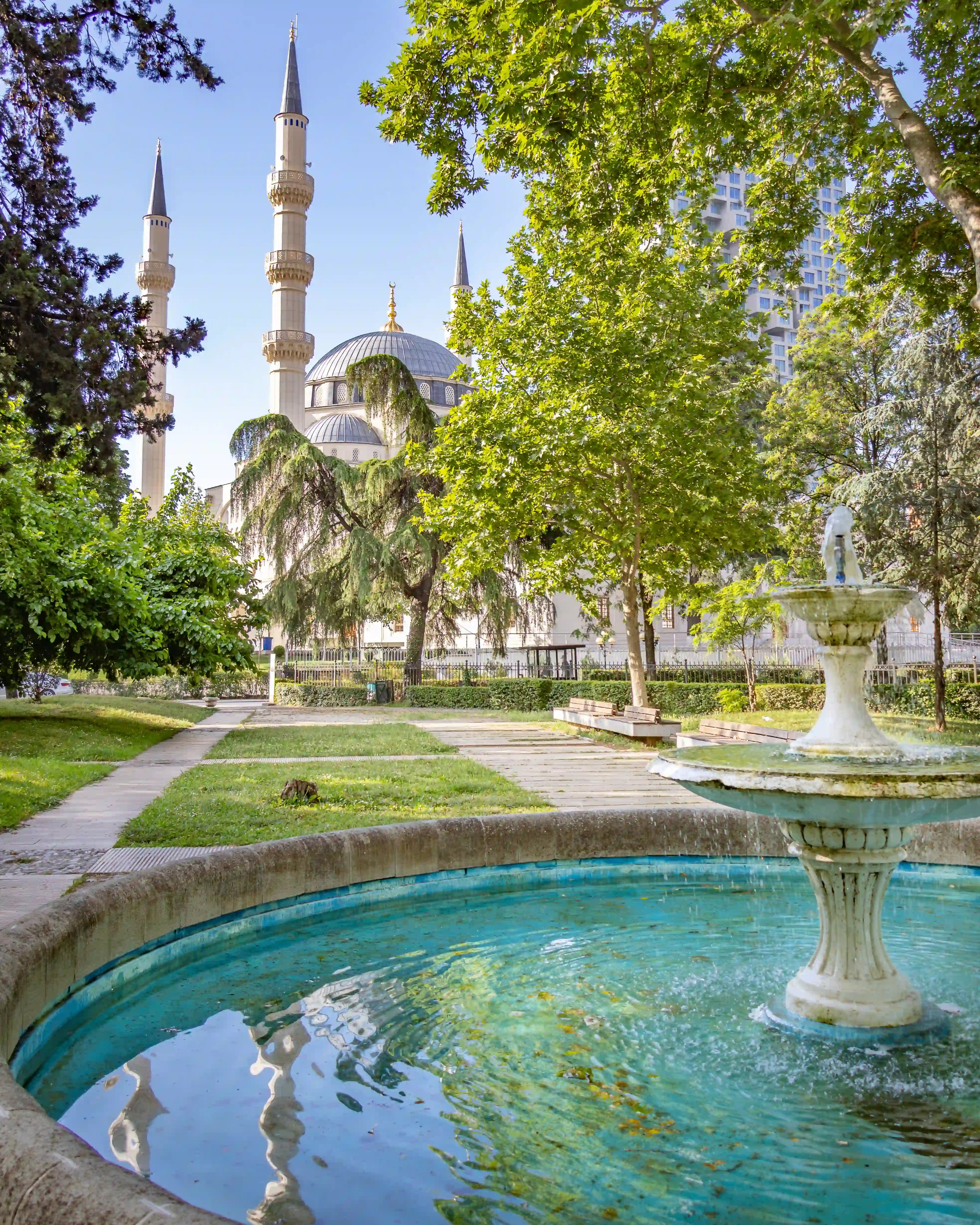 Fountain in the gardens near the Grand Mosque of Tirana with minarets visible through the trees.