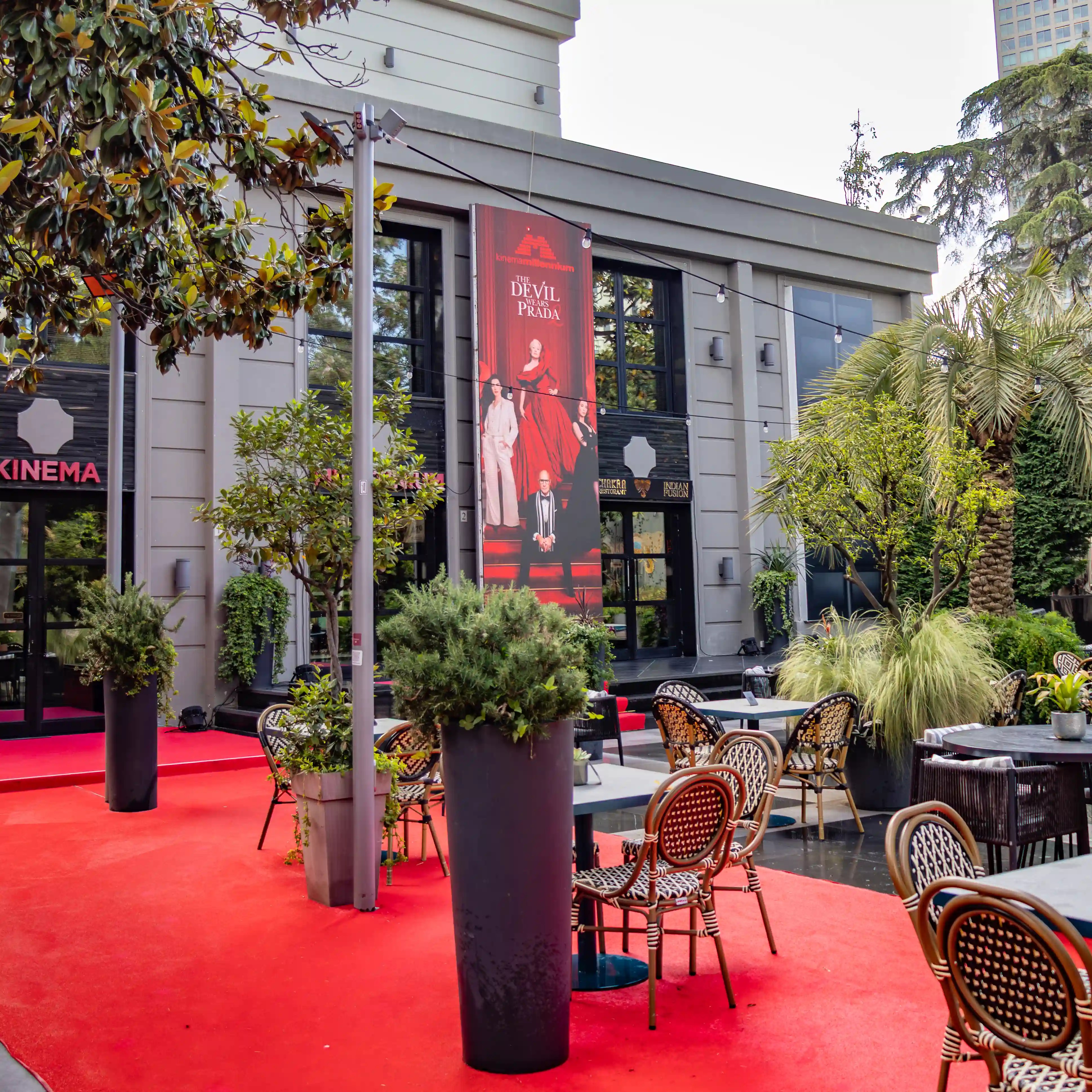 An outdoor courtyard with café tables, potted plants, and a red carpet leading toward a modern cinema building in Tirana.