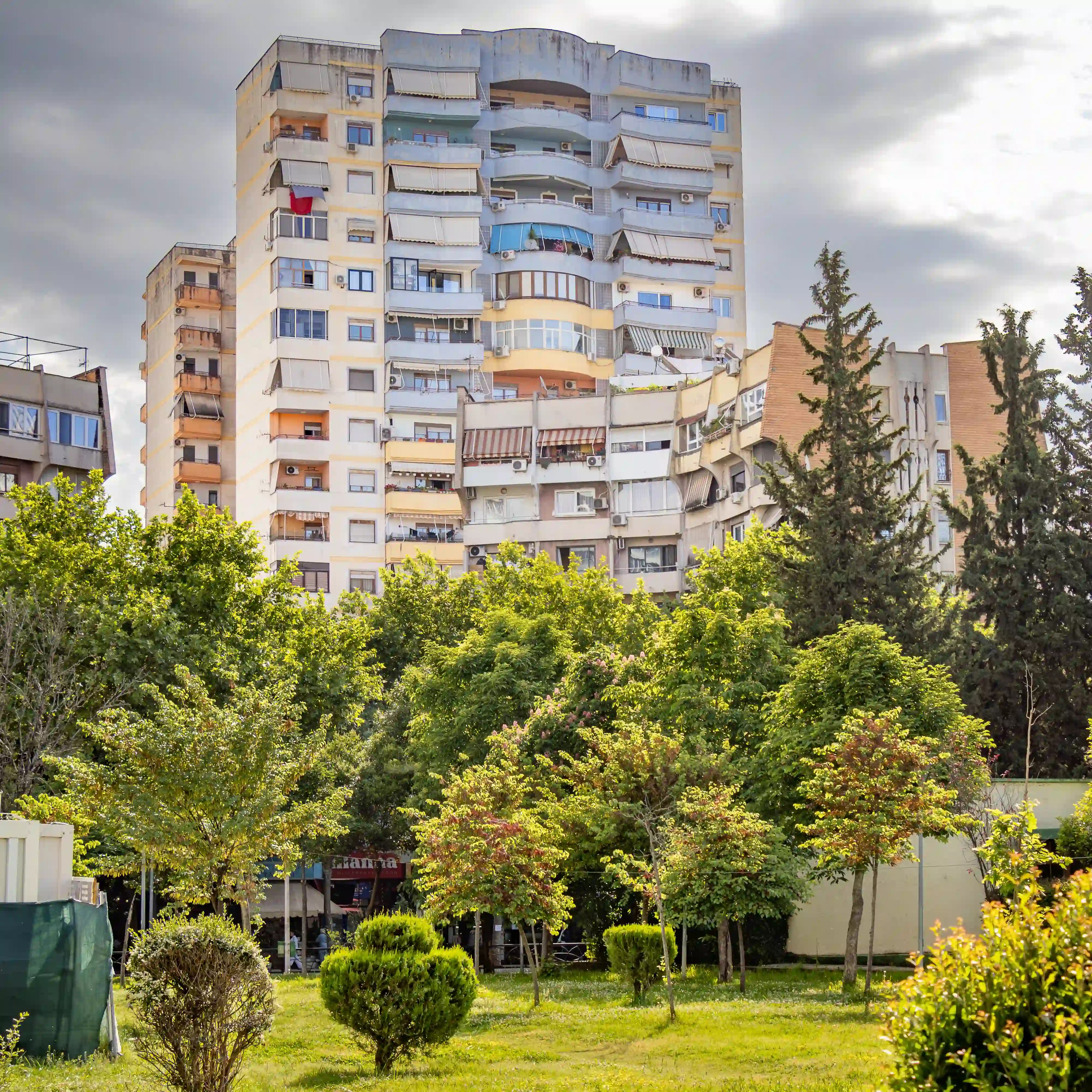 Communist-era apartment buildings rise above a green park filled with trees under a cloudy sky in Tirana.