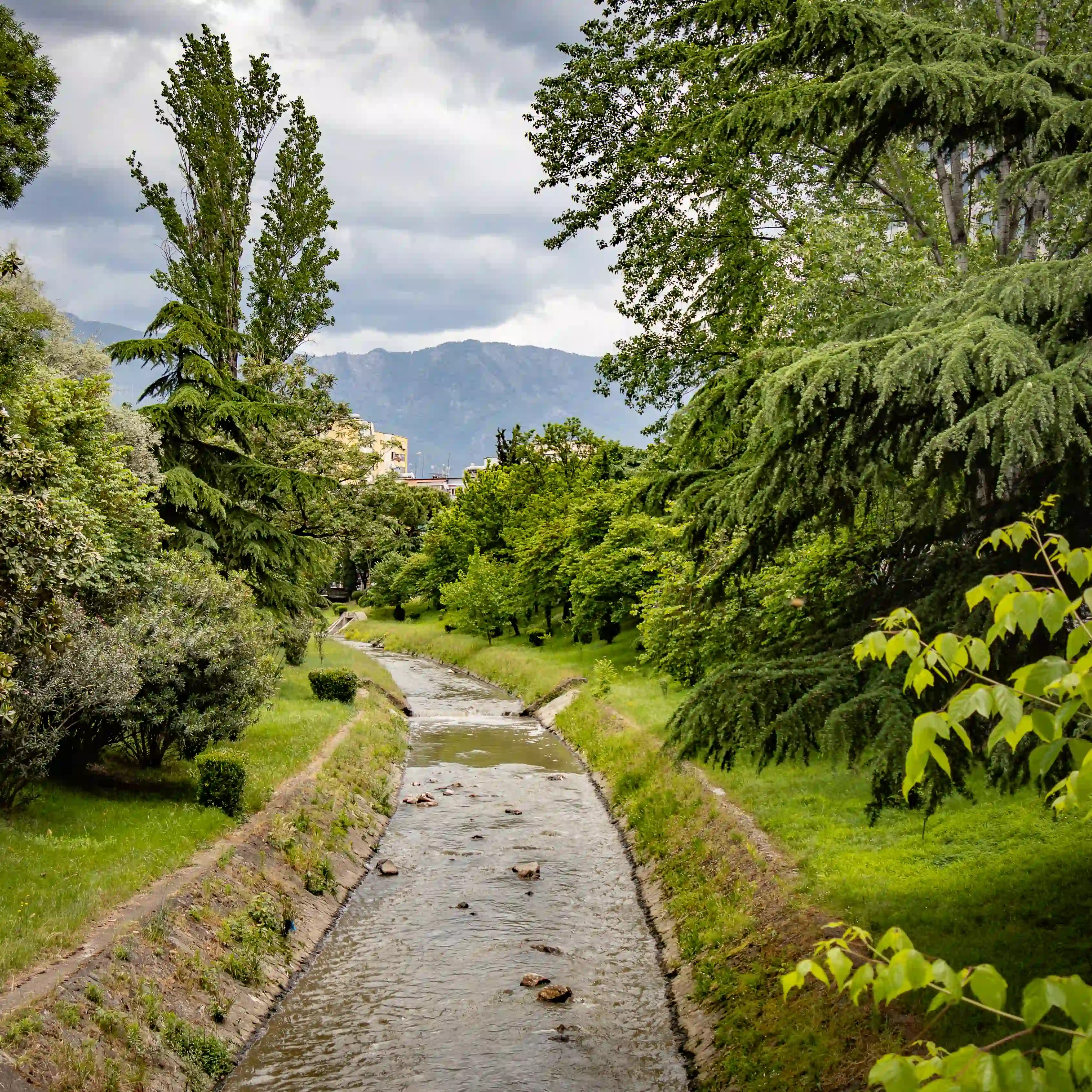 A narrow canal lined with trees and grass flows through a green park with mountains visible in the distance.