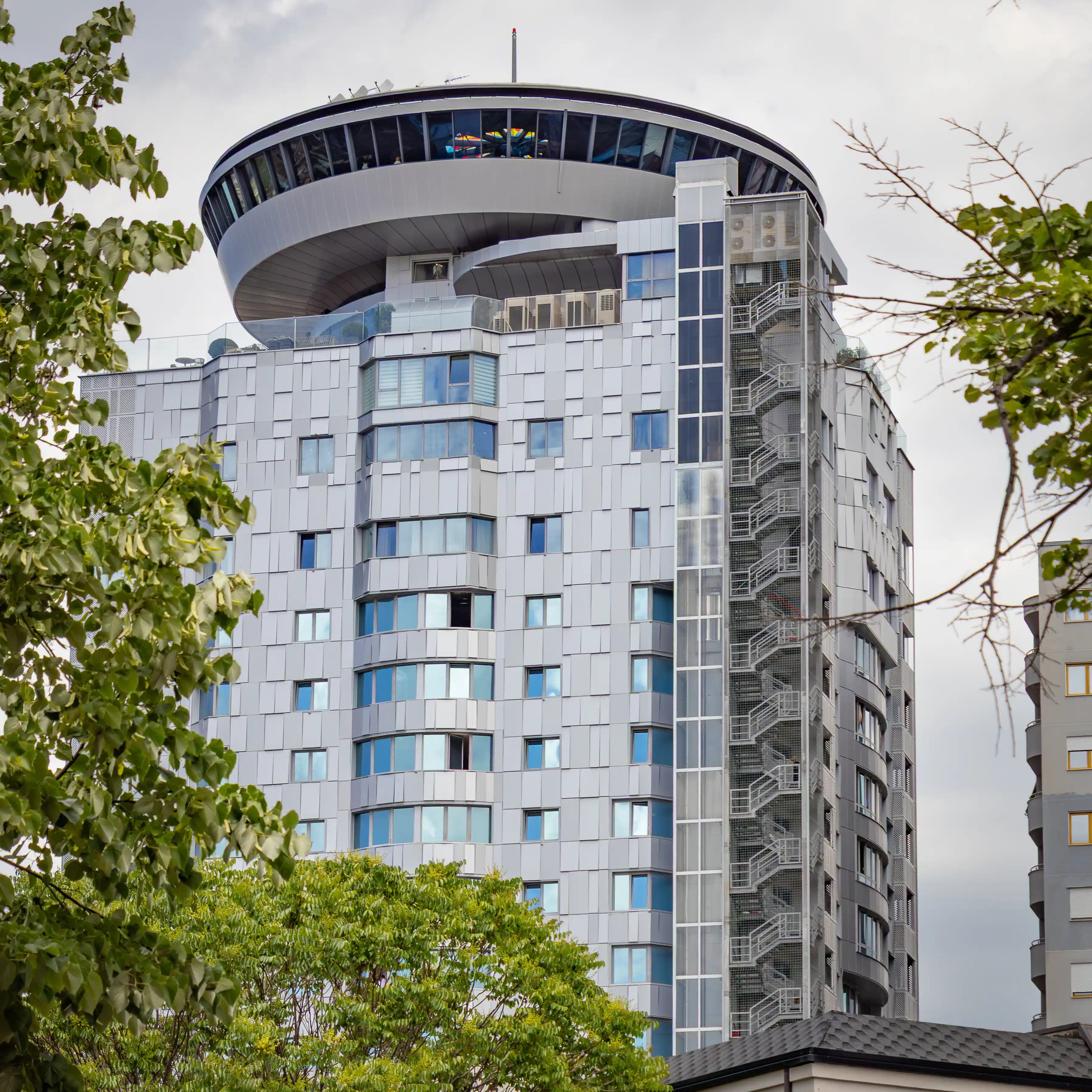 A modern high-rise building with a circular rooftop structure rises above trees in Tirana.