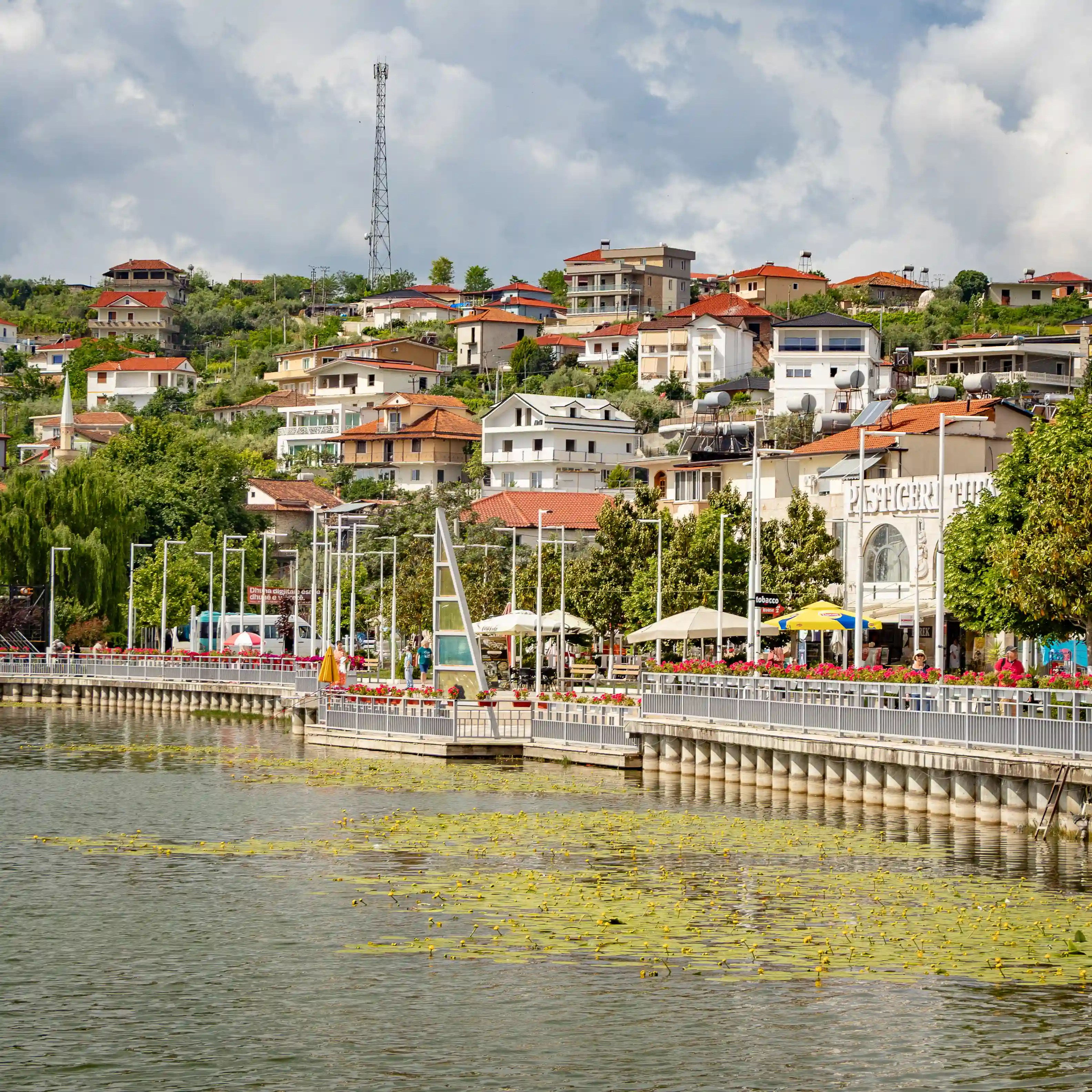 A lakeside promenade lined with flowers and cafés curves around Belshi Lake with hillside homes in the background.