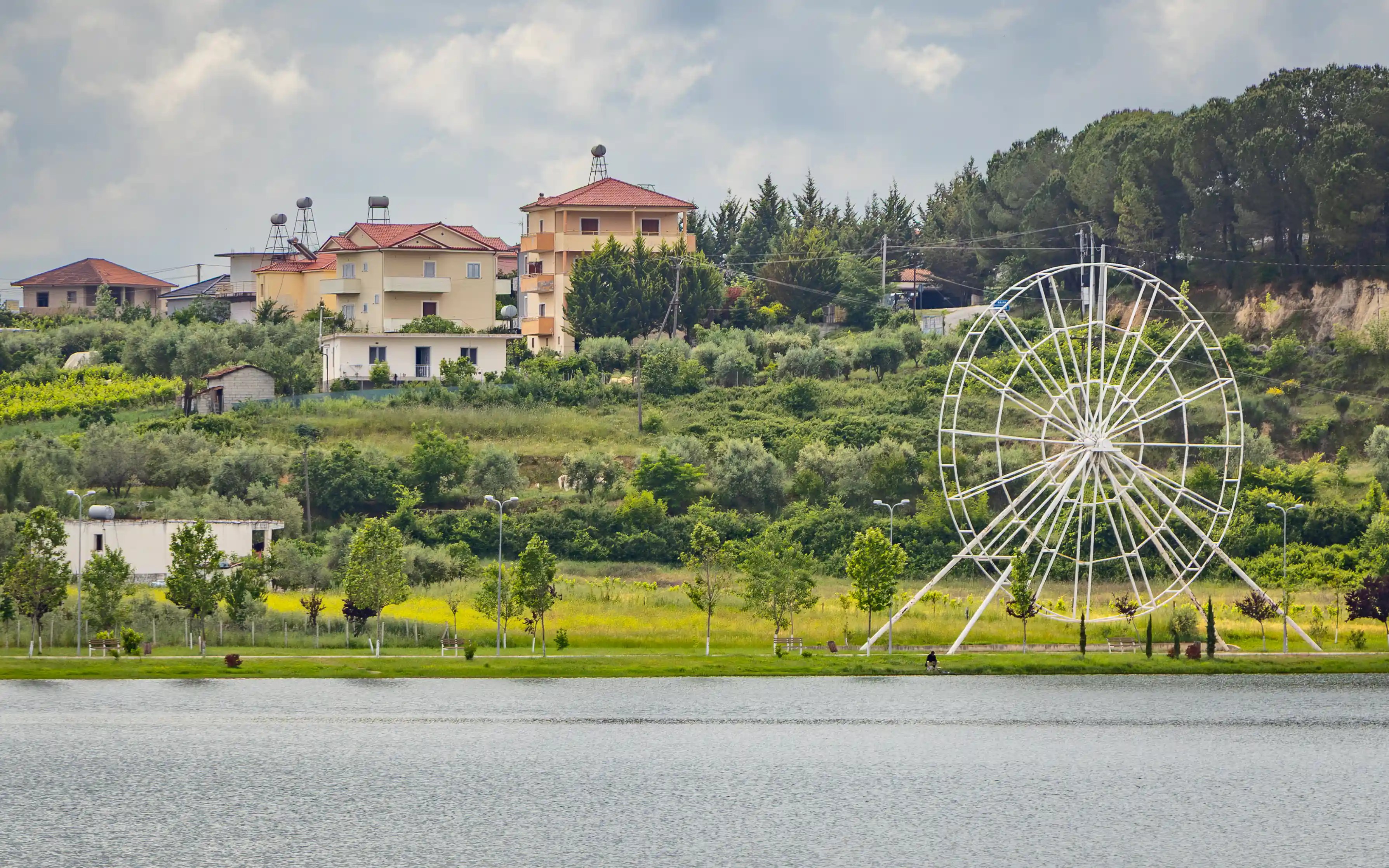 A white Ferris wheel stands near Belshi Lake with homes and green hills in the background.