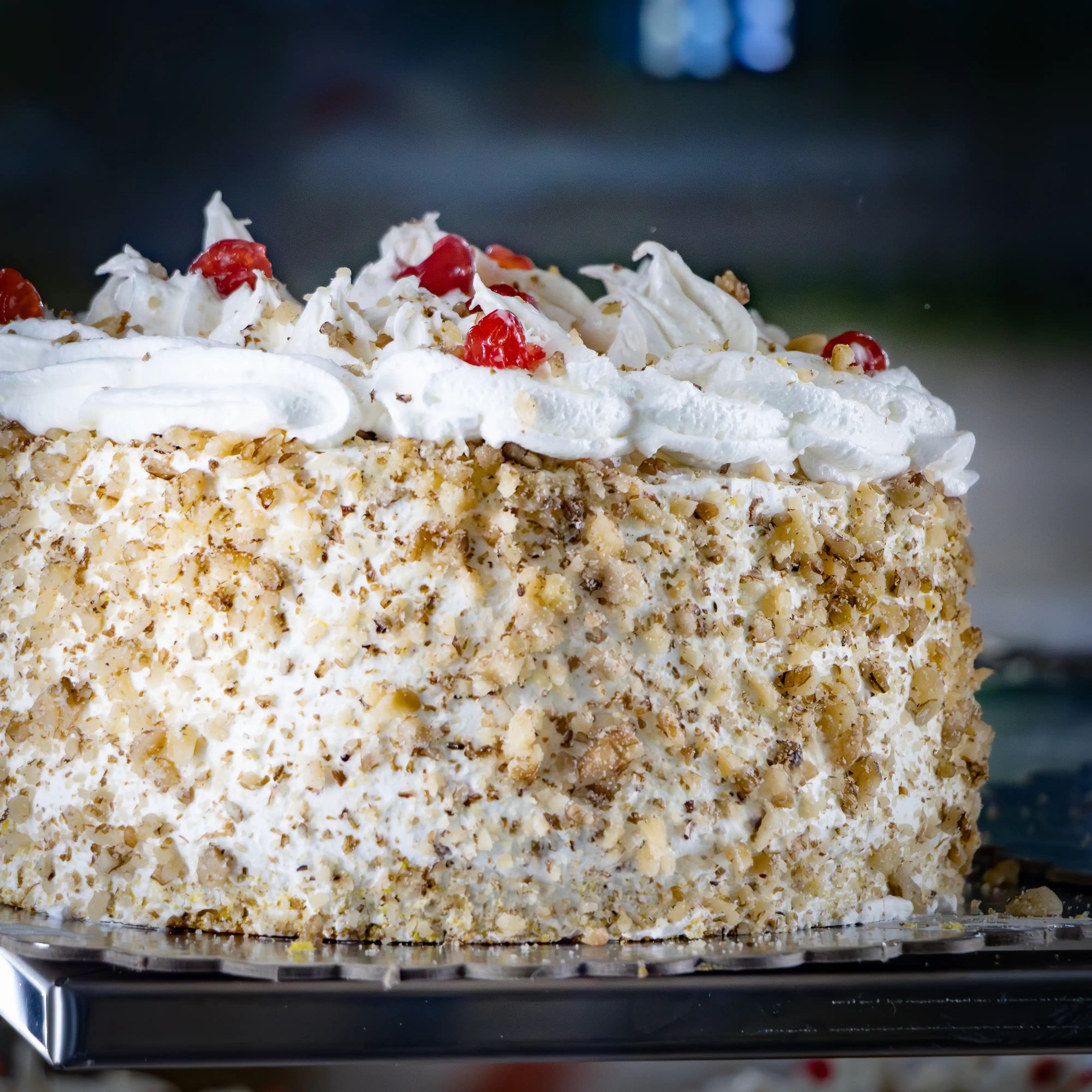 A cream-covered cake topped with walnuts, whipped cream, and cherries sits on display in a bakery case.