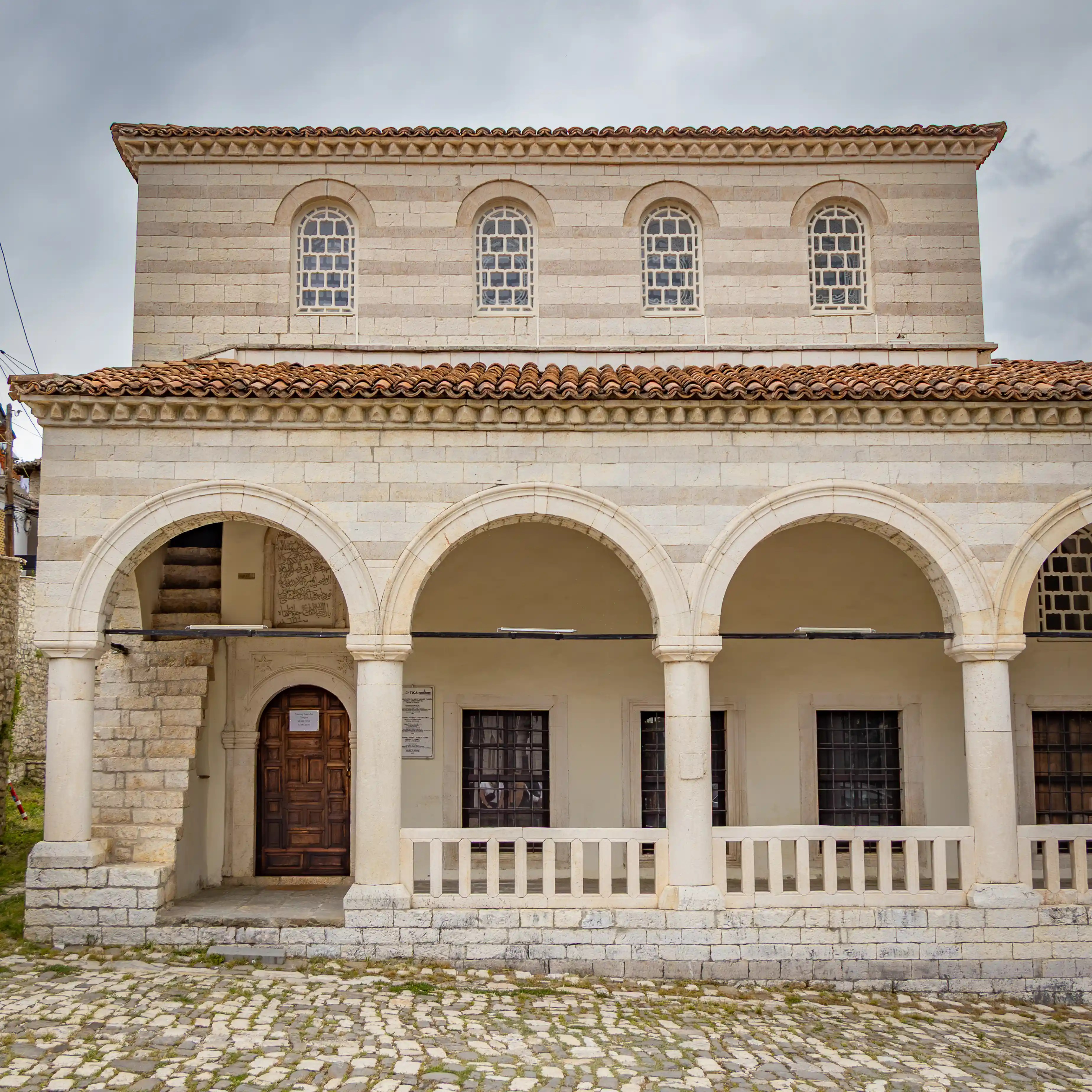 The historic Helveti Tekke with arched arcades and a tiled roof stands along a cobblestone street in Berat.
