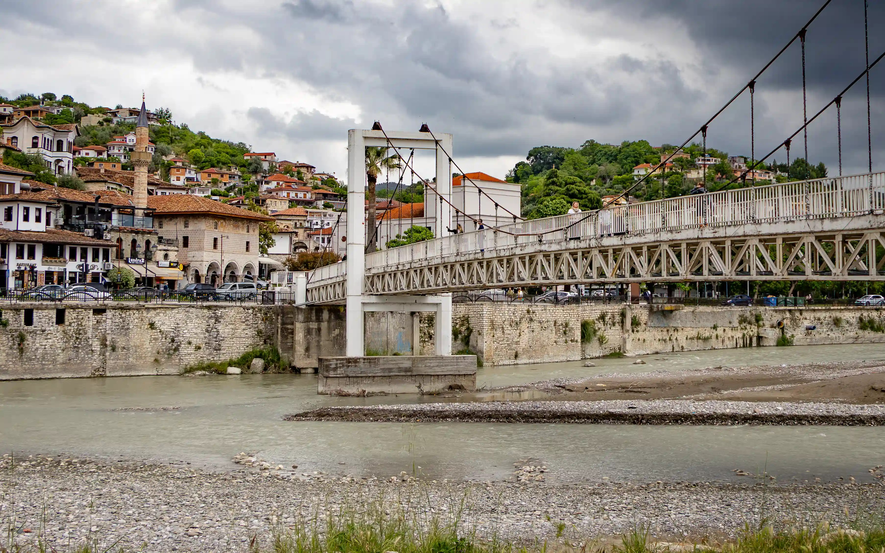 The white Suspension Bridge crosses the Osum River in Berat beneath dark clouds.