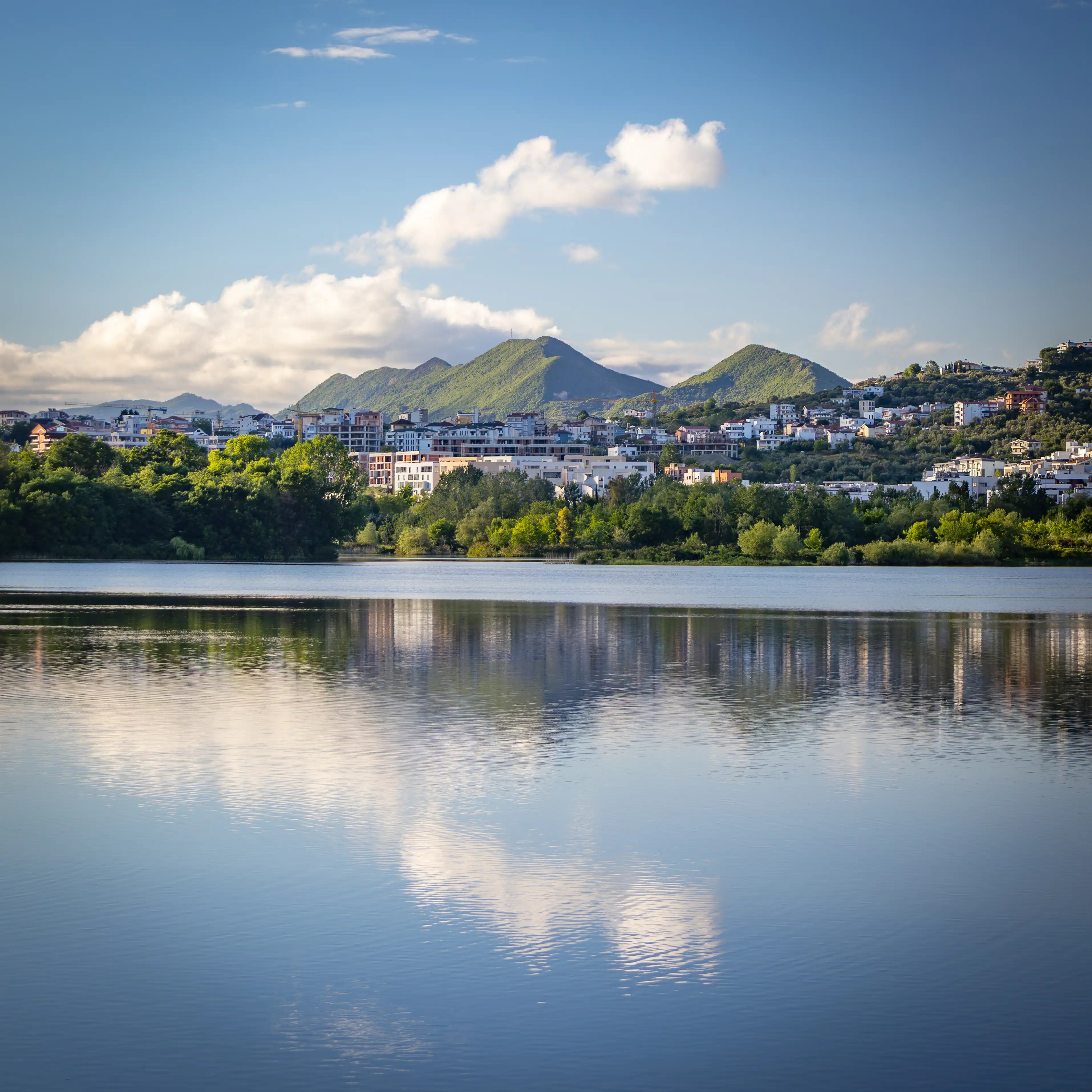 View across Tirana's Artificial Lake toward green hills and neighborhoods reflected in the calm water.