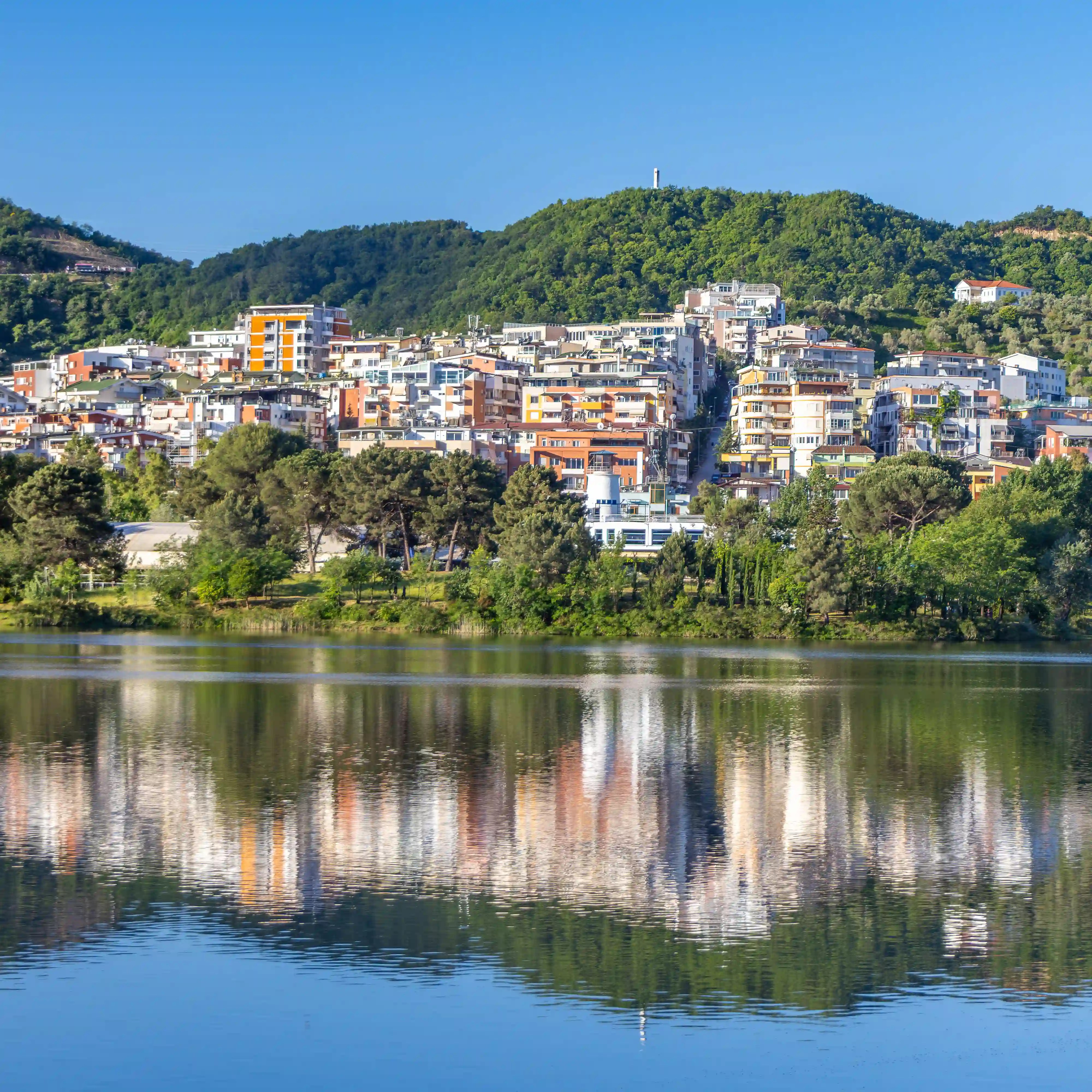 Residential buildings on a hillside reflected in the calm waters of Tirana's Artificial Lake.