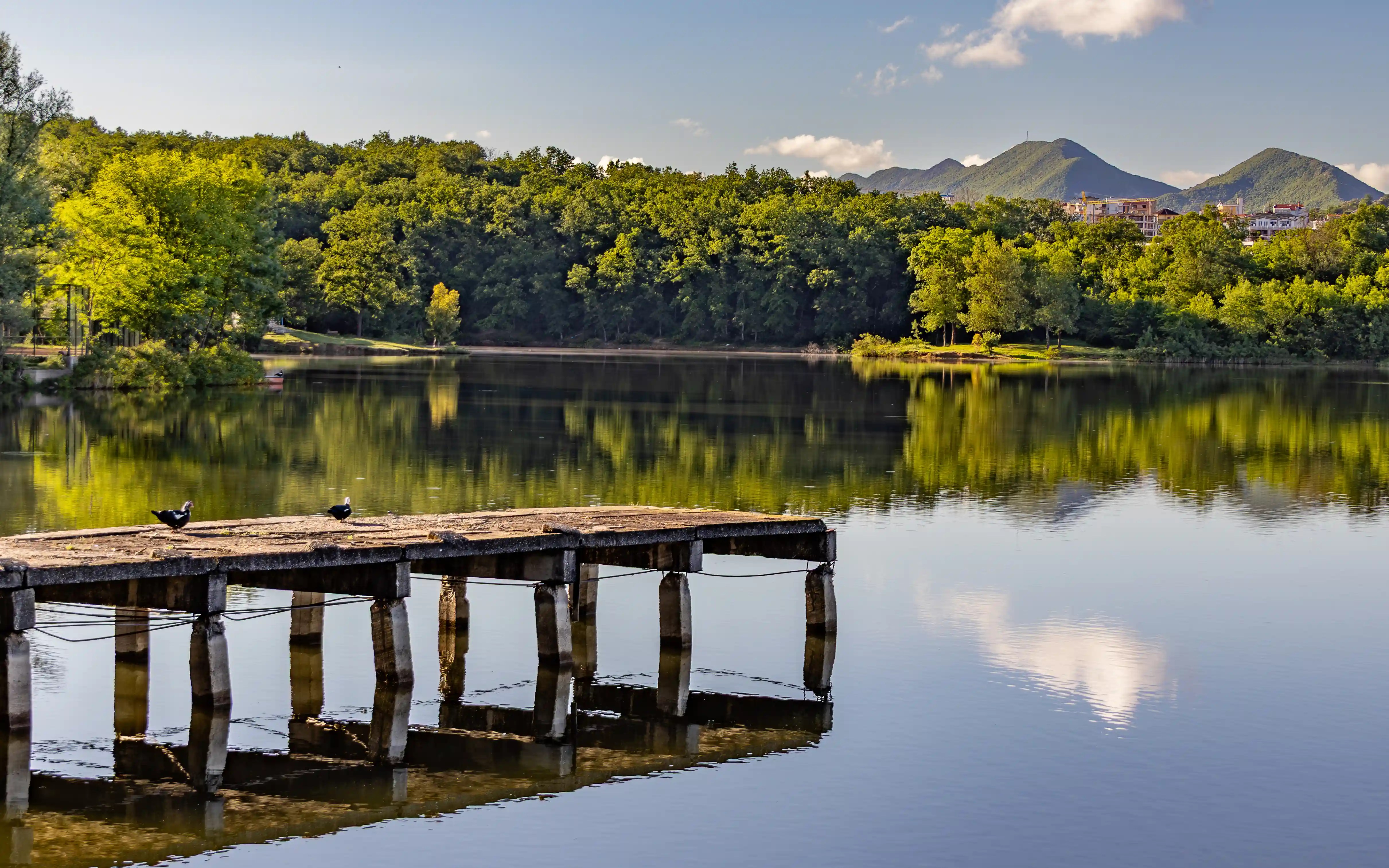A small concrete pier extending into Tirana's Artificial Lake with hills and trees reflected in the water.