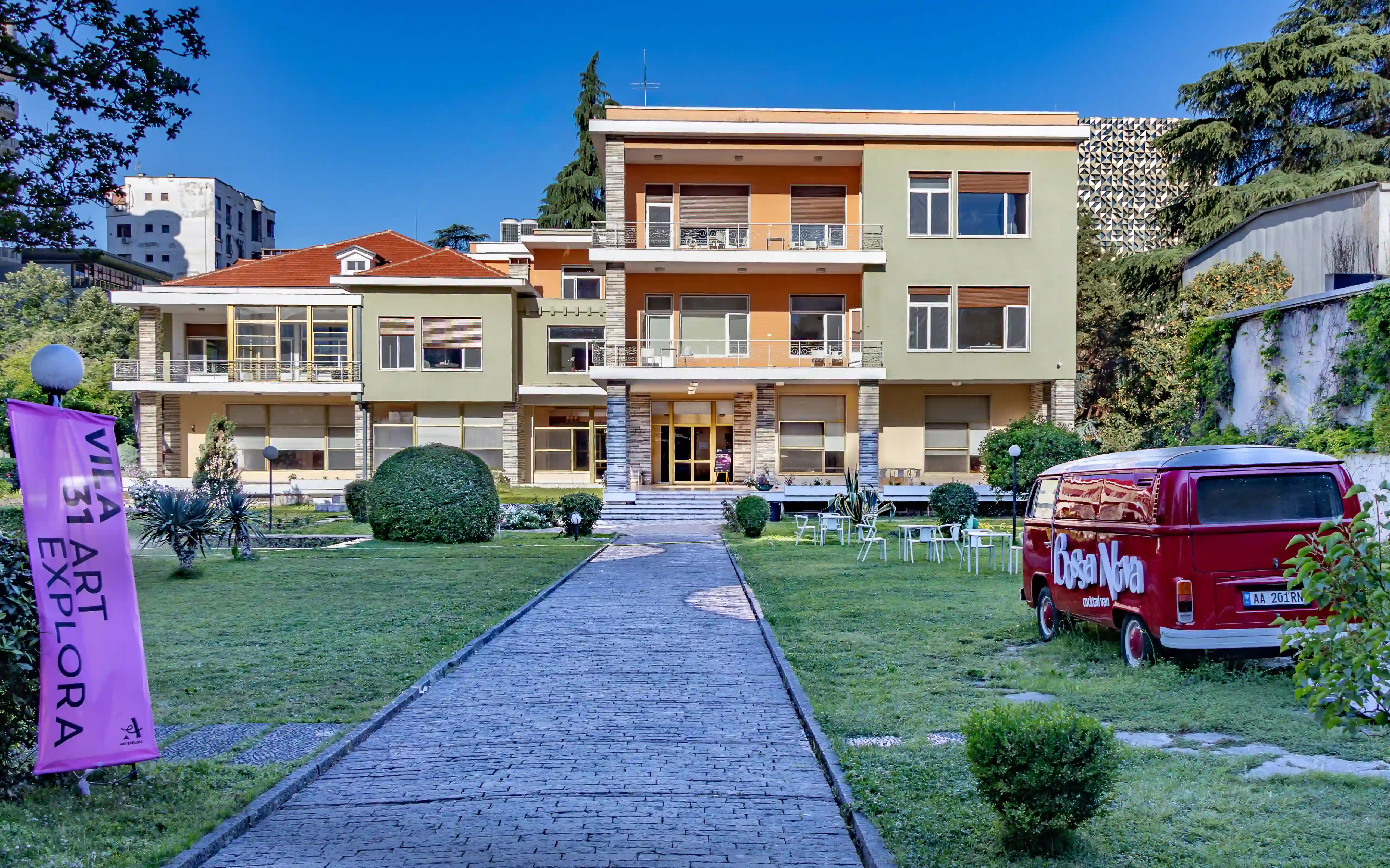 The former home of Enver Hoxha in Tirana, a large villa with landscaped grounds, outdoor seating, and a vintage red van parked on the lawn.