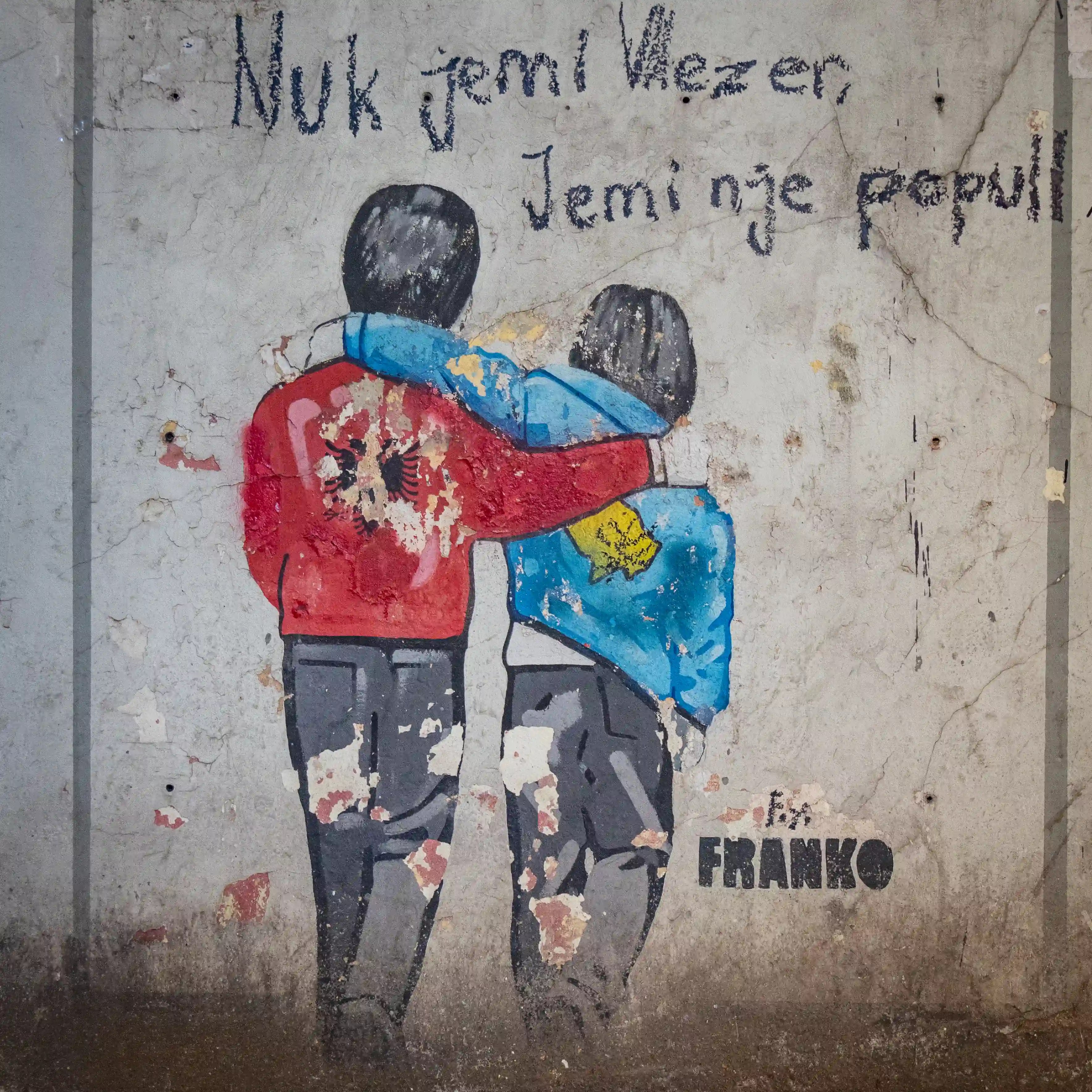 A weathered mural shows two children walking arm in arm, draped in Albanian and Kosovo flags, painted on a concrete wall.